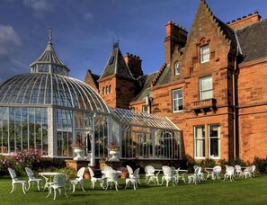 A large glass conservatory with a dome roof, surrounded by white metal chairs and tables, set against a brick building with a steeply pitched roof.