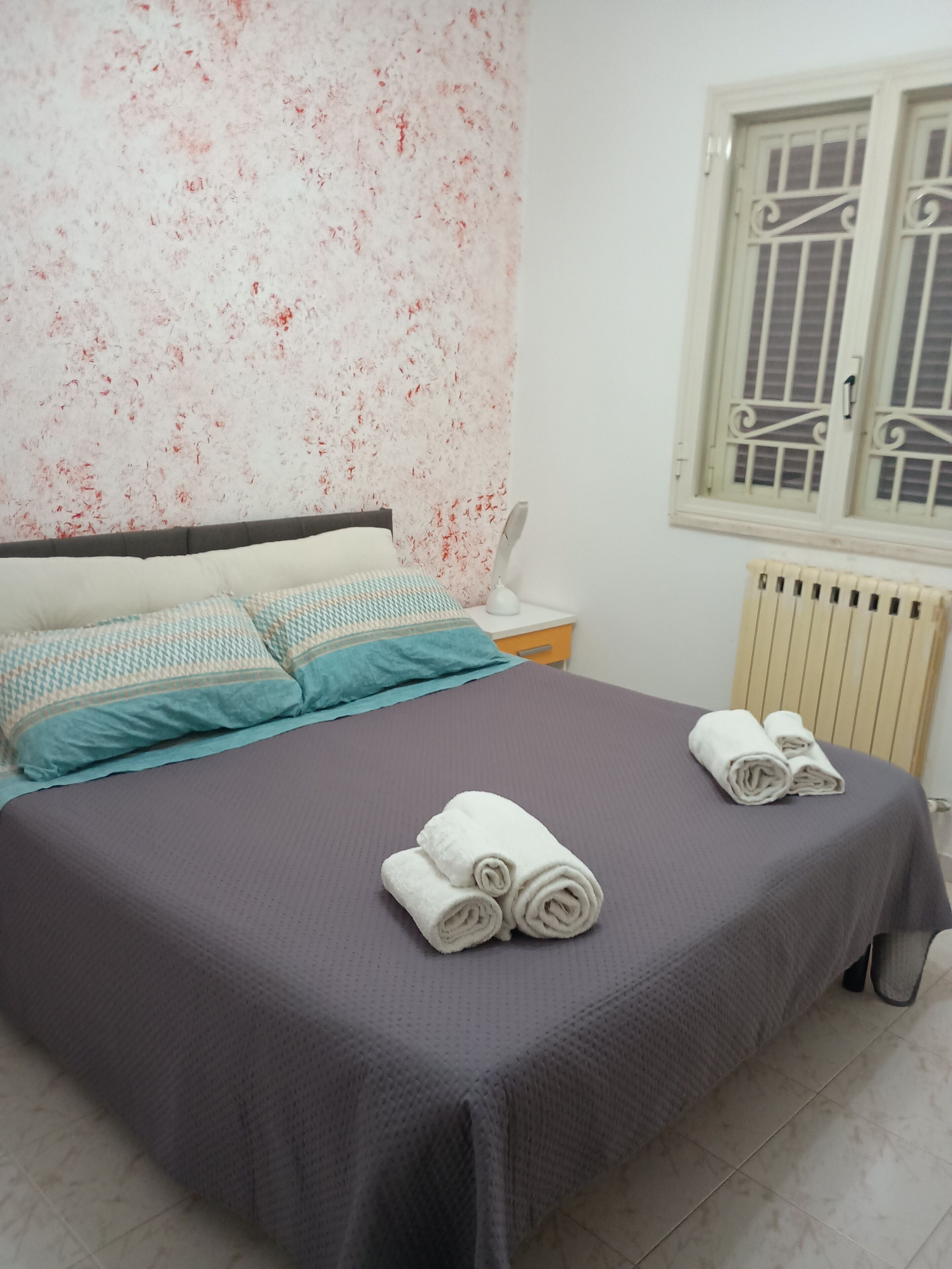 A neatly made bed with a grey bedspread and blue pillows, a white bedside table with a lamp, and a window with decorative bars.