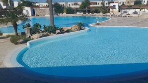 Una grande piscina a forma di curva con lettini e ombrelloni, circondata da edifici e palme.