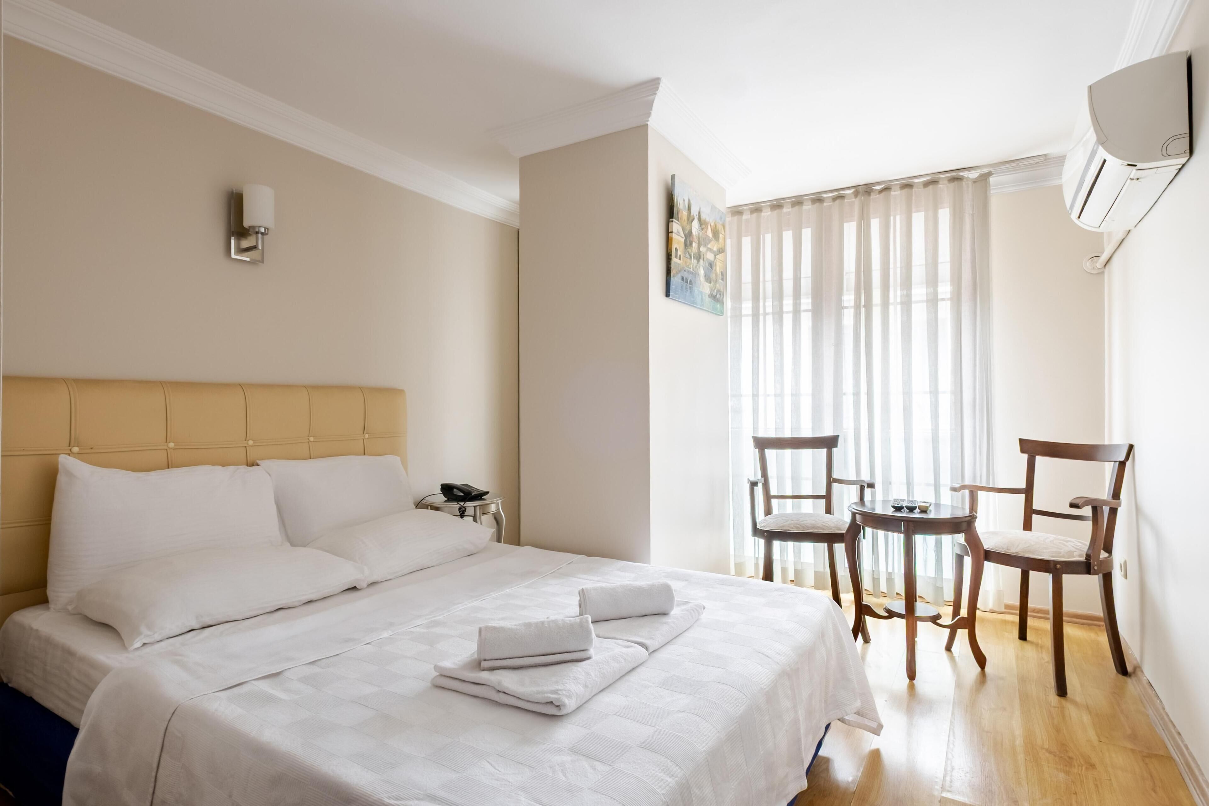 A hotel room with a bed, a desk, two chairs, a painting, and a window with curtains.