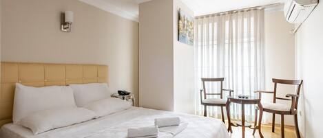 A hotel room with a bed, a desk, two chairs, a painting, and a window with curtains.