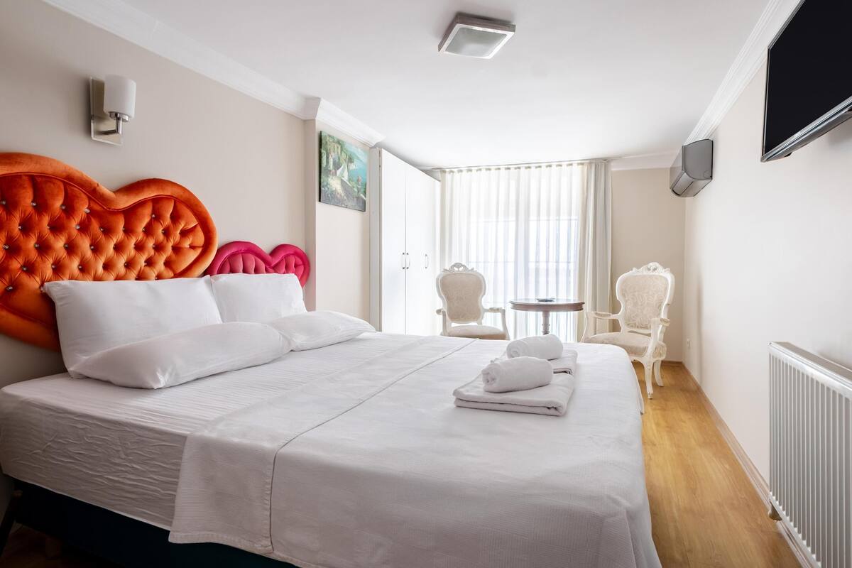 A hotel room with a large bed, a TV, a window with blinds, and a chair.