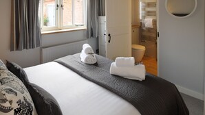 A bedroom with a large bed, a window with curtains, a bathroom visible through an open door, and a round mirror on the wall. - Swallows Nest cottage sleeps 10 guests in 6 bedrooms. (Fakenham)