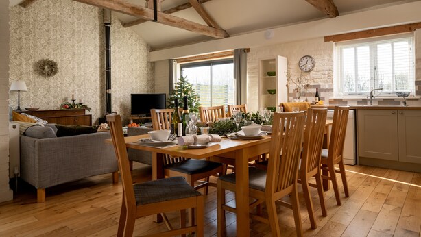 A dining area with a wooden table and chairs, a television, a fireplace, and a kitchen in the background. - Swallows Nest cottage sleeps 10 guests in 6 bedrooms. (Fakenham)