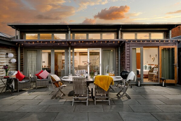 A modern house with a patio area featuring outdoor furniture, a dining table, and a view of the interior. - Eco Barn - a communicating cottage that sleeps 12 guests in 6 bedrooms (Fakenham)