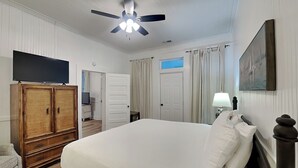 Une chambre d’hôtel avec un lit, un téléviseur posé sur une commode en bois, un ventilateur de plafond, une fenêtre avec des rideaux, une porte et un tableau accroché au mur.