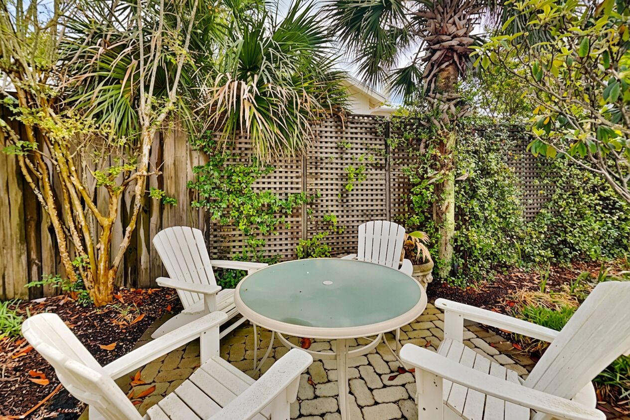 A patio with a round table and four white chairs, surrounded by a wooden fence and various plants.