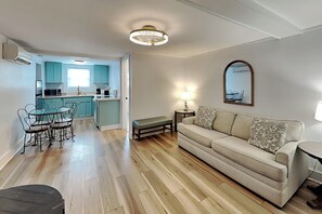 Ein Wohnzimmer mit einer Couch, einem Esstisch mit Stühlen, einer Küchenzeile mit türkisfarbenen Schränken und einer Deckenleuchte.