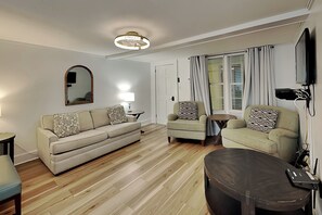 Ein Wohnzimmer mit einer Couch, Sesseln, einem Couchtisch, einem Spiegel, einer Lampe, einem Fernseher und einem Fenster mit Vorhängen.