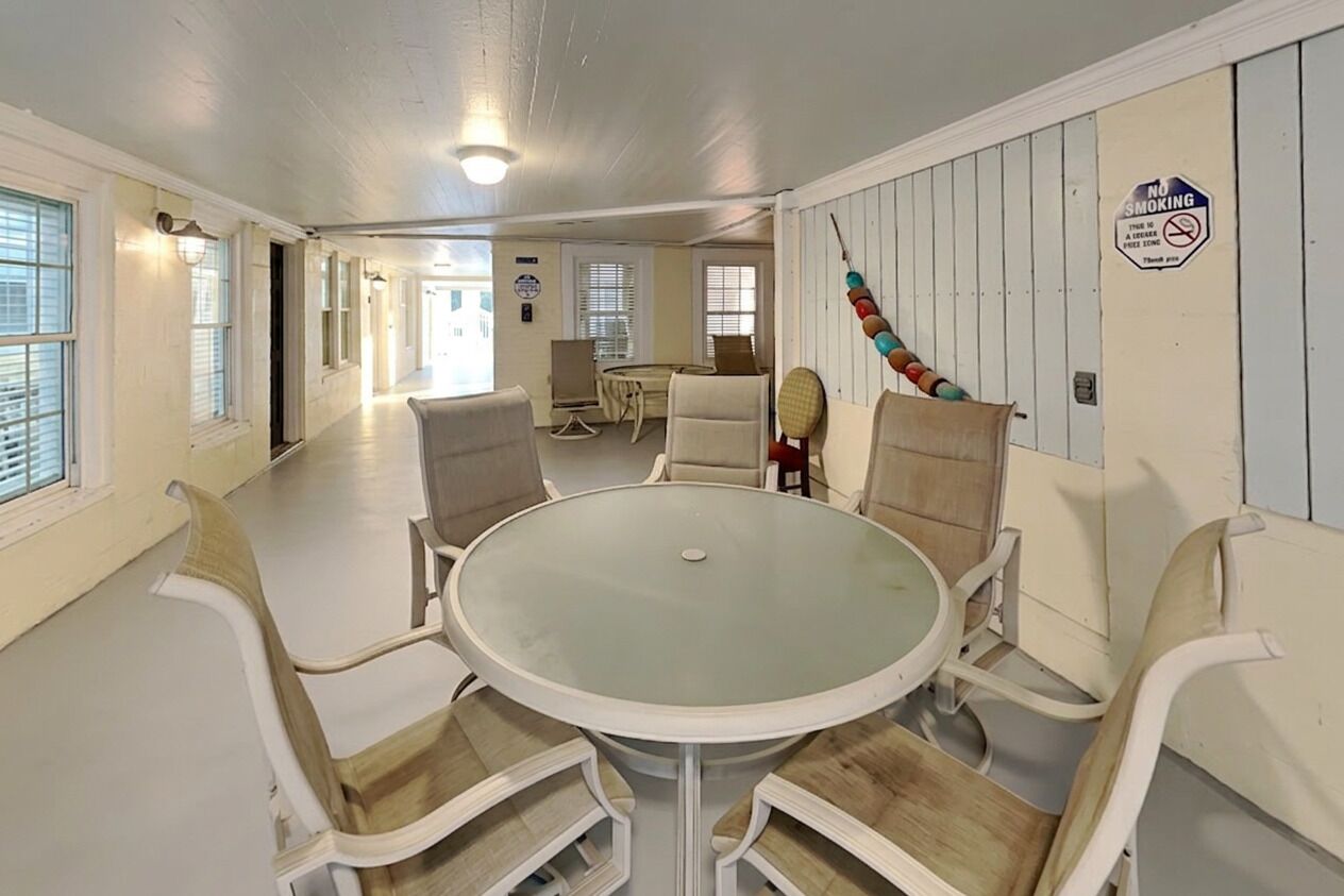 A dining area with a round glass table and white chairs, a no-smoking sign, and a view of the adjacent room.