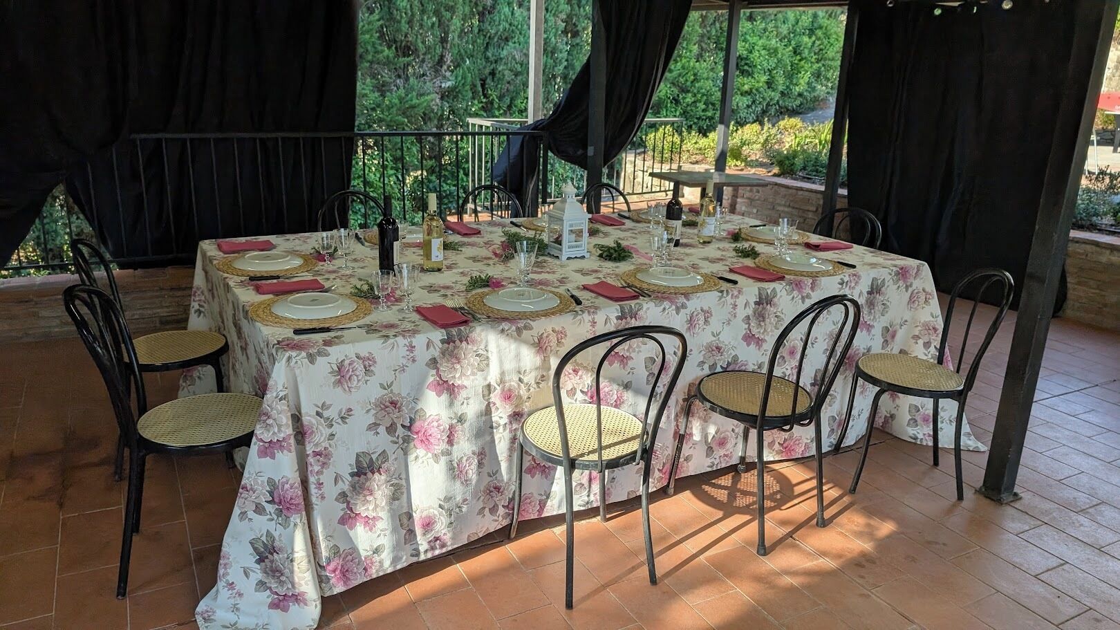 An outdoor dining area with a table set for four, surrounded by chairs, under a covered patio with a view of greenery.