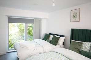 A bedroom with a bed, a green headboard, a window with a view of greenery, and a framed picture on the wall. - Stylish 2 bed home near Trafford  (Eccles)