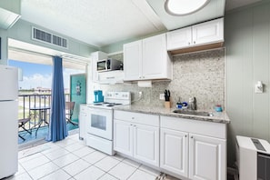 A kitchen with white cabinets, a microwave, stove, and a sink. There is a balcony with a blue curtain and a view of the outside.