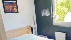 Une chambre à coucher avec un lit, une fenêtre donnant sur un paysage verdoyant, un radiateur et deux tableaux encadrés accrochés au mur.