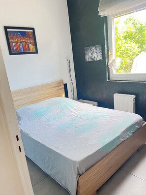 A bedroom with a bed, a window with a view of greenery, a radiator, and two framed pictures on the wall. - Le clos des mimosas (Bormes-les-Mimosas)