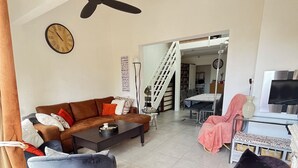 Un salon avec un canapé marron, un fauteuil bleu, un fauteuil violet, une table basse, un ventilateur de plafond, une horloge et un escalier menant à une autre pièce.