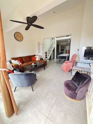 A living room with a brown sofa, a blue armchair, a purple armchair, a coffee table, a ceiling fan, a clock, and a staircase leading to another room. - Le clos des mimosas (Bormes-les-Mimosas)