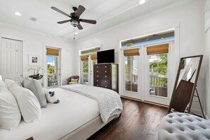 Ein Schlafzimmer mit Bett, Deckenventilator, Kommode, Fernseher und großen Fenstern mit Jalousien.