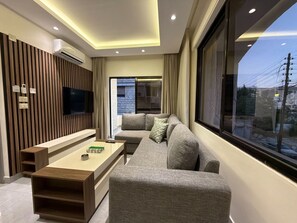 Un salon moderne comprenant un canapé, une table basse, un téléviseur fixé sur un mur en panneaux de bois, et une grande fenêtre donnant sur des immeubles.