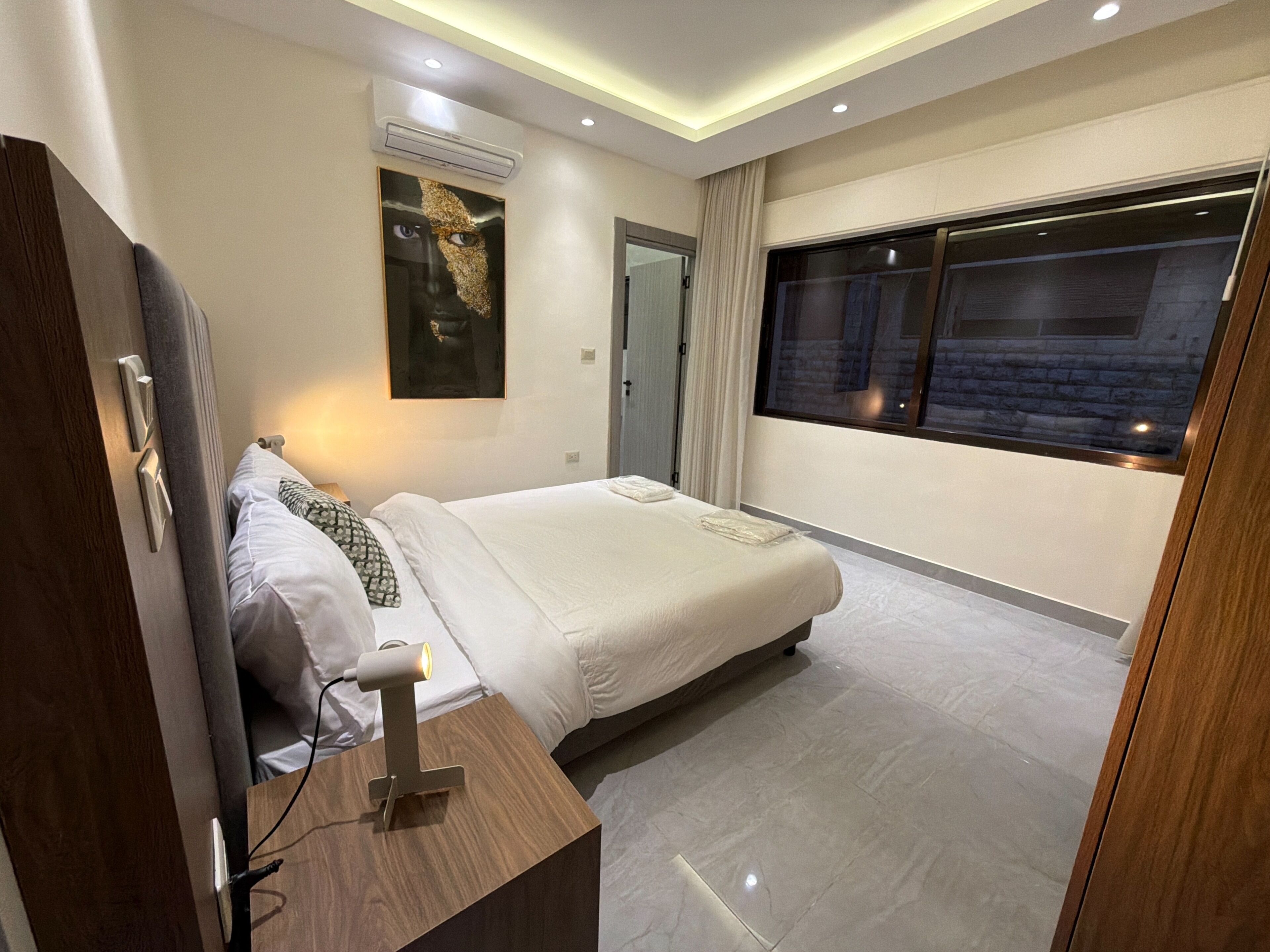 Ein modernes Hotelzimmer mit Bett, Nachttisch mit Lampe, Fenster mit Blick und Klimaanlage.