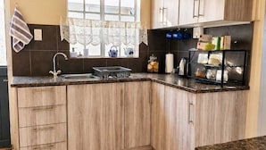 A kitchen with wooden cabinets, a sink, a window with lace curtains, and a countertop with kitchen items. - Amazing 2-bedroom cottage in Graafwater, perfect for a relaxing getaway (Graafwater)
