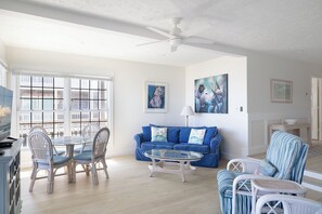 Ein Wohnzimmer mit blauem Sofa, einem Glastisch, Korbstühlen und einem Deckenventilator.