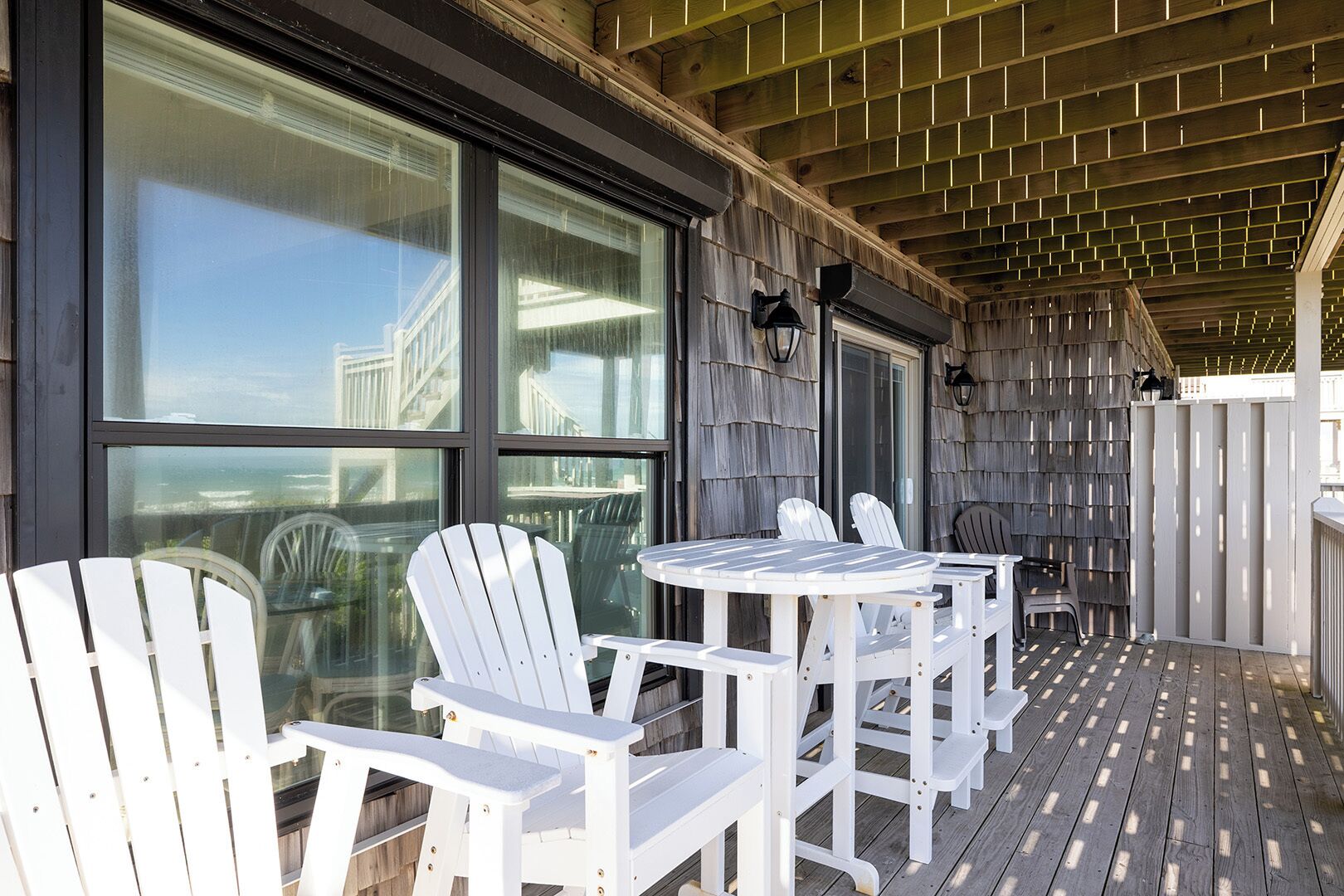 A covered outdoor seating area with Adirondack chairs and a table, large windows, and a wooden deck.