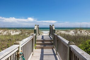 Ein hölzerner Steg führt zu einem Strand mit Dünen und wolkenlosem, blauem Himmel.