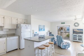 Eine Küche mit weißen Schränken, einem Kühlschrank, einem Essbereich mit gestreiften Stühlen und Blick auf das Meer durch eine Tür.