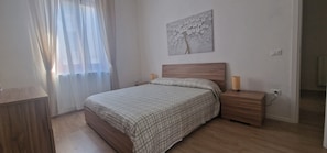 Une chambre à coucher comprenant un lit, une table de chevet, une lampe, un tableau décoratif et une fenêtre avec des rideaux.