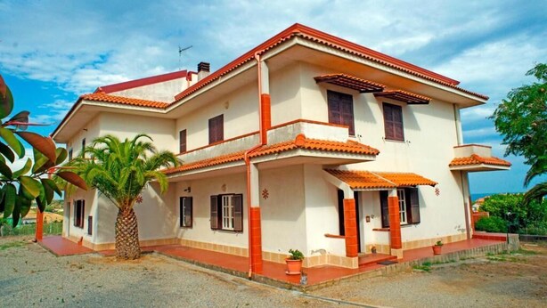 Una casa de dos pisos con techo de tejas rojas, un porche cubierto y una palmera en primer plano.