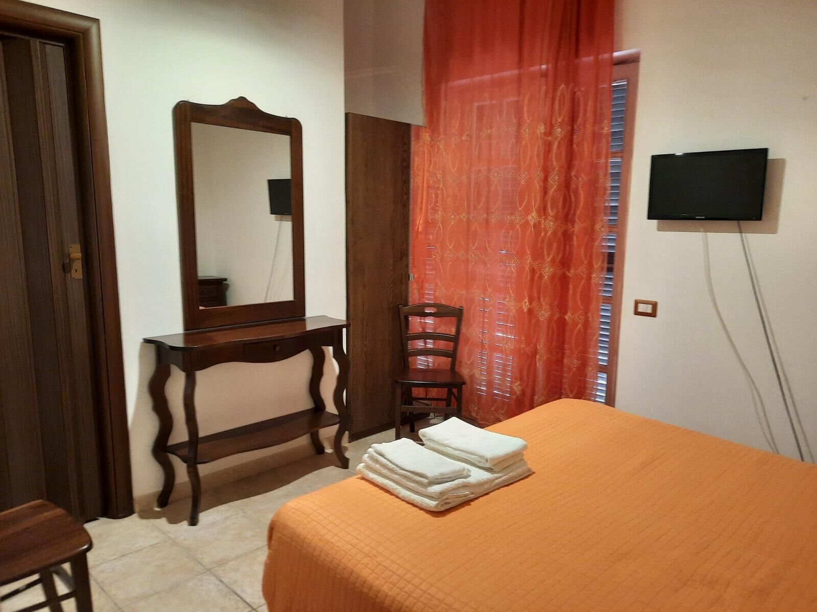 Een slaapkamer met een bed, een houten dressoir, een stoel, een aan de muur bevestigde televisie en een raam met gordijnen.