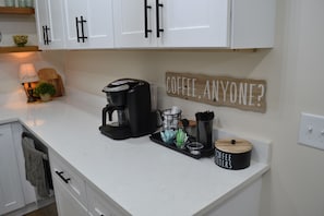 A kitchen with white cabinets, a coffee maker, a coffee filter container, and a sign that reads 'COFFEE, ANYONE?'