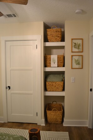 A hallway with a white door, wicker baskets on shelves, and framed botanical prints on the wall.