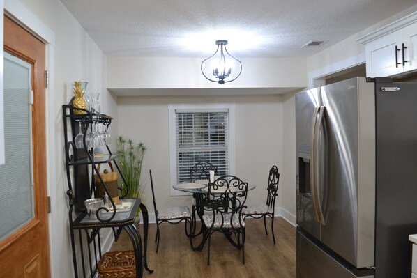 A kitchen with a stainless steel refrigerator, a dining table with four chairs, a chandelier, and a window with blinds.