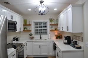 A modern kitchen with white cabinets, stainless steel appliances, a window with blinds, and a coffee station.