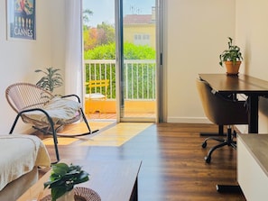 Ein Zimmer mit Holzboden, einem Korbstuhl, einem Schreibtisch, einem Topfpflanzen und einer Glastür zum Balkon mit gelbem Stuhl und Pflanzen.