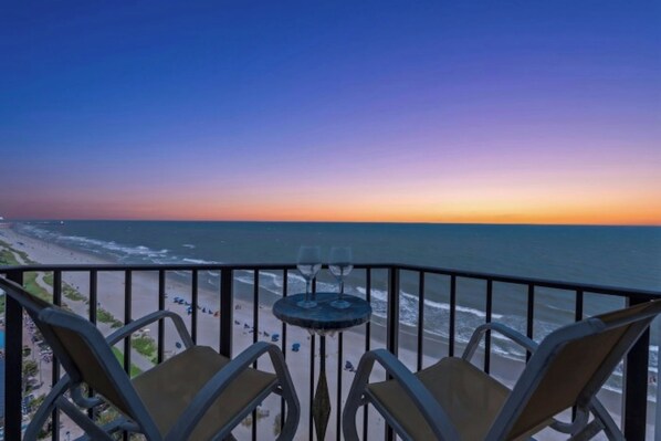 Ein Balkon mit zwei Stühlen und einem kleinen Tisch mit Blick auf einen Strand und das Meer bei Sonnenuntergang.