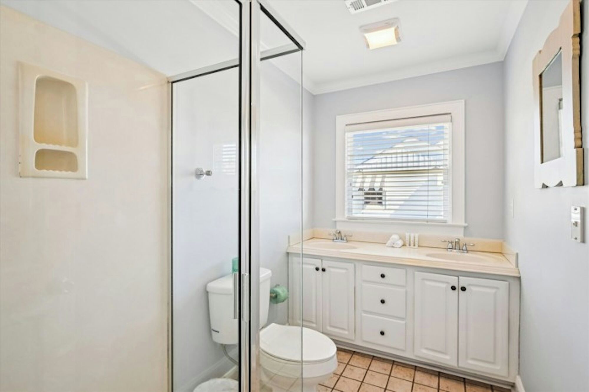 Un baño con cabina de ducha de vidrio, inodoro, lavabo con espejo y ventana con persianas.