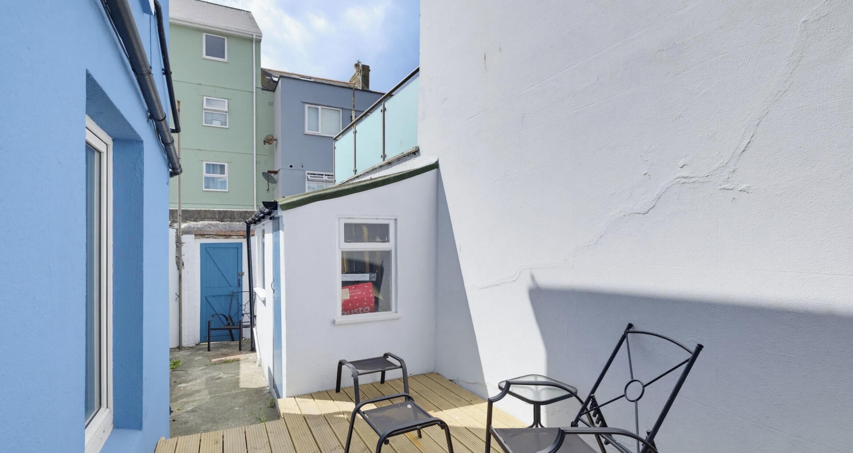 A narrow alleyway with a blue building on one side and a white building on the other. There are two metal chairs and a small table in the foreground.