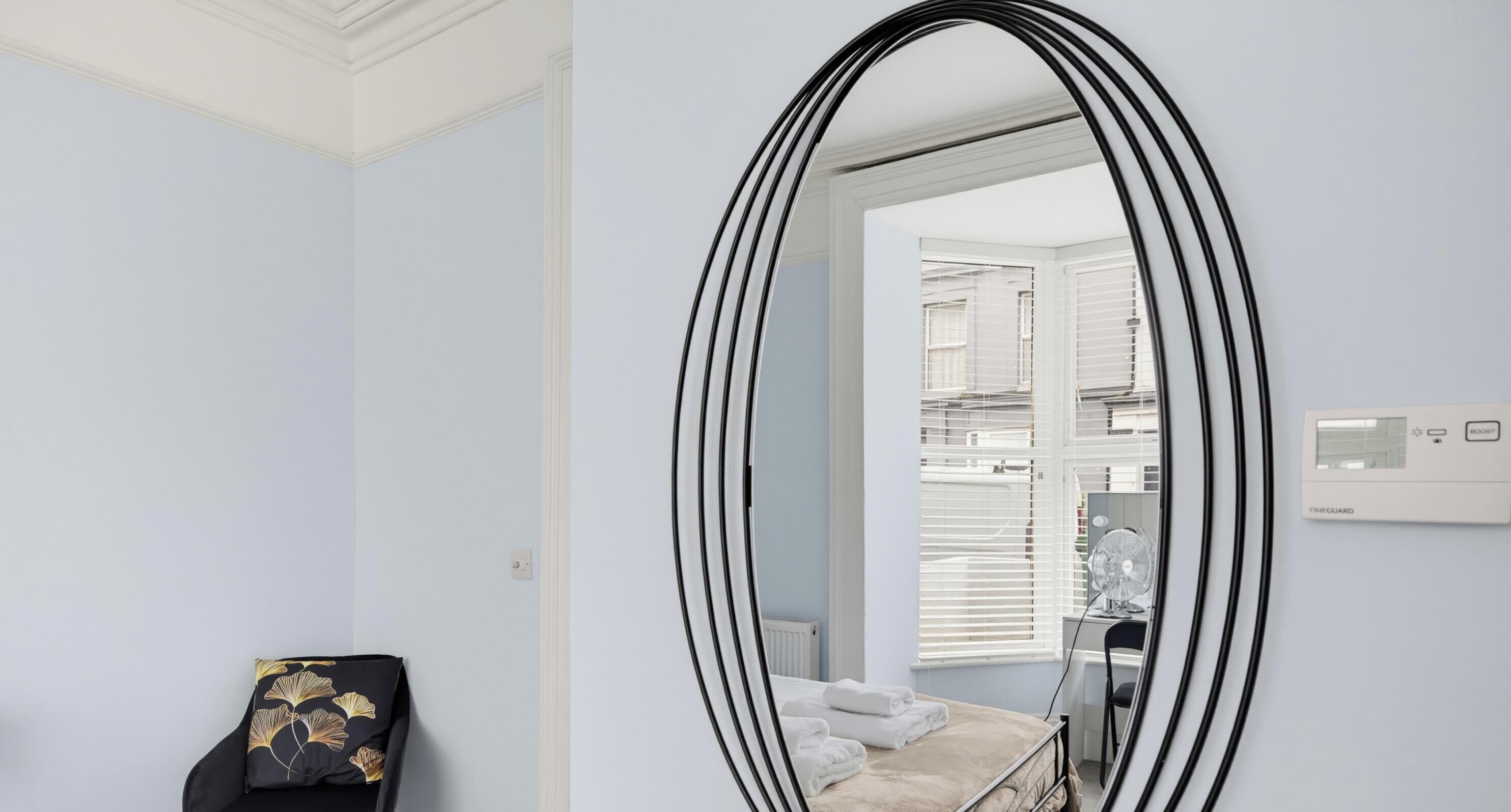 Una habitación con un espejo ovalado grande, una cama con ropa blanca, un sillón con cojín negro y una ventana con persianas.