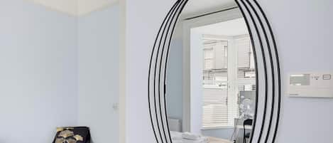 Una habitación con un espejo ovalado grande, una cama con ropa blanca, un sillón con cojín negro y una ventana con persianas.