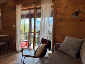 Interior d'una cabana de fusta amb taula de dinar, una cadira amb almoçadet, un sofà i un finestral gran amb cortines.