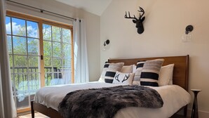 Een slaapkamer met een groot bed, een opgehangen hertenkop, een lamp van een glazen fles en uitzicht door een raam.