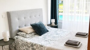 A bedroom with a grey upholstered headboard, a floral-patterned bedspread, a black rug, a bedside table with a lamp, and a window with curtains. - Bed and breakfast with shared pool (10m2) (GODINHAÇOS)