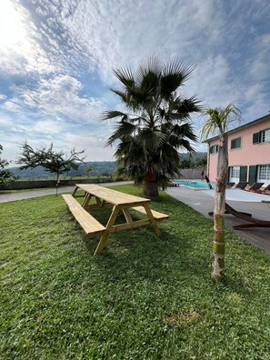 A picnic table on a grassy lawn with a palm tree and a building with windows in the background. - Bed and breakfast with shared pool (10m2) (Braga)