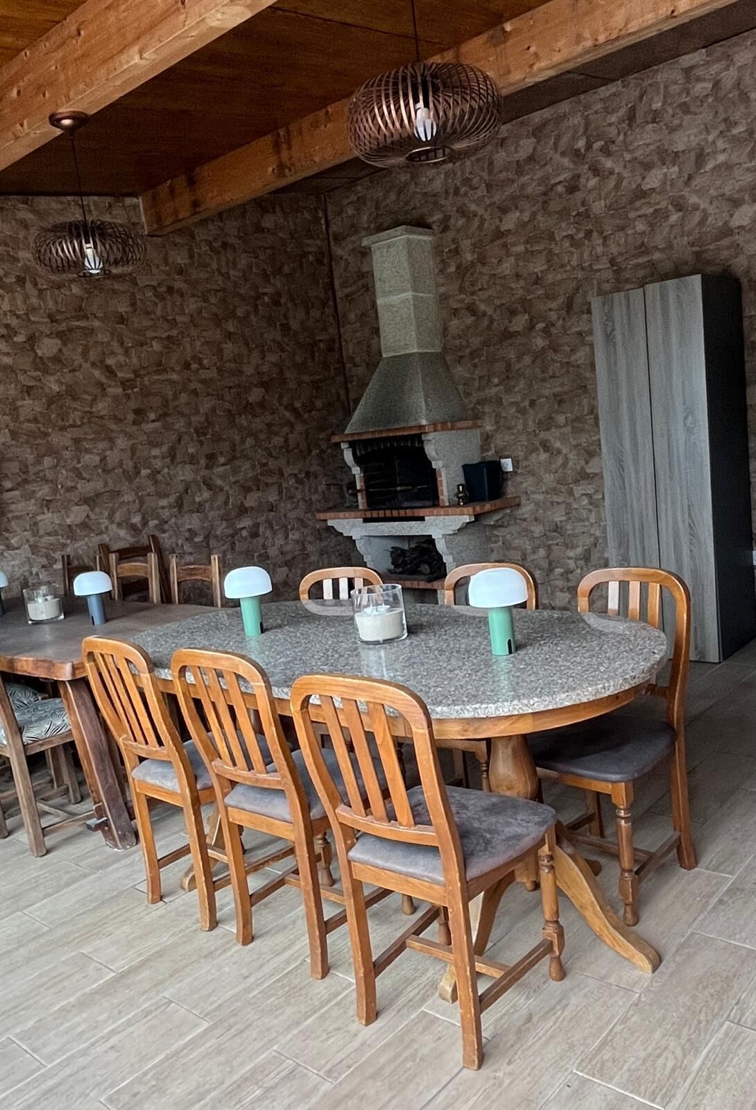 A rustic dining area with a stone wall, a chimney, and wooden chairs and tables.