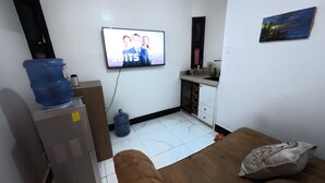 A small living area with a sofa, a TV displaying a show, a water dispenser, and a kitchenette.