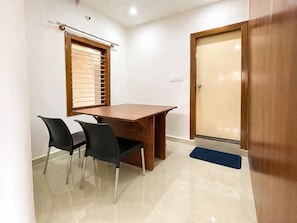 A room with a wooden table, two black chairs, a window with blinds, a door, and a blue mat.