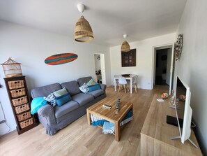 Ein Wohnzimmer mit grauem Sofa, Holztisch, einer Hängeleuchte aus Weidwerk und einem Essbereich mit weißem Tisch und Stühlen.